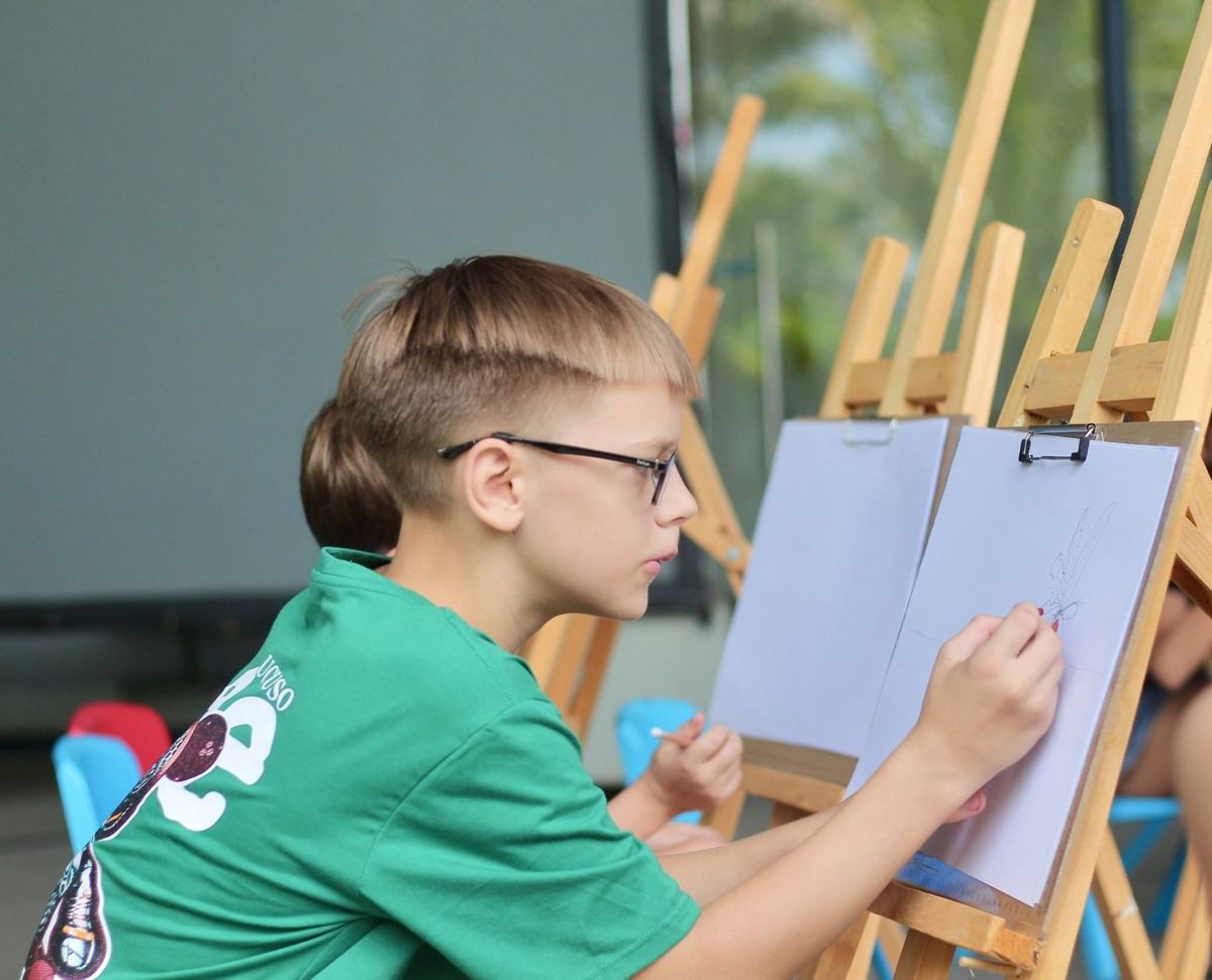 Boy in glasses sketching at a wooden easel in a painting class